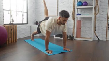 Spor salonunda mavi bir minder üzerinde yoga yapan genç İspanyol bir adam, kapalı bir spor merkezinde egzersiz teknikleri gösteriyor..