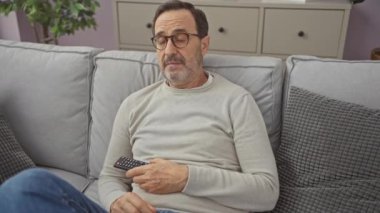 Olgun adam evdeki bir kanepede oturup günlük rahatlama tasviri yaparken uzaktan kumandayı tutuyor..