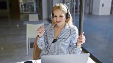 Kulaklık takan neşeli sarışın bir kadın modern bir ofis masasında laptopuyla çalışıyor..