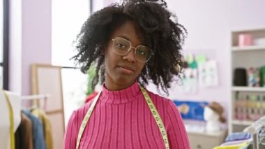 Afro-Amerikalı bir kadın, mankenler ve kumaşlar görünür bir terzi dükkanında güvenle gülümsüyor..