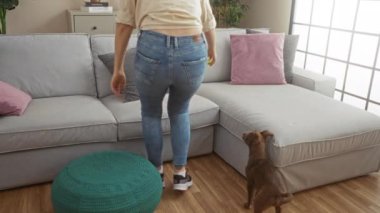 Genç bir kadın köpeğiyle rahat bir oturma odasında oynuyor. İçinde bej bir kanepe, renkli yastıklar ve deniz mavisi bir puf var..