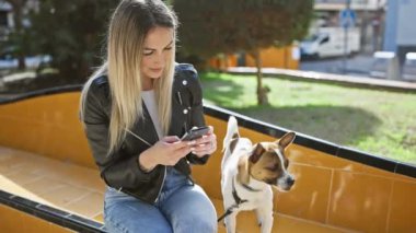 Sarışın kadın parktaki bankta dikkatli köpeğinin yanında akıllı telefon kullanıyor..