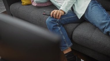 Genç Asyalı bir adam rahat bir oturma odasında, gri bir koltukta, renkli yastıklarla, uzaktan kumandayla rahatlar..