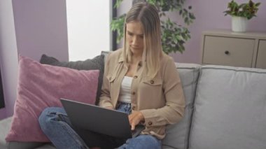 Evindeki oturma odasında oturan hayal kırıklığına uğramış genç bir kadın kanepede dizüstü bilgisayar kullanıyor. Yüzünde endişeli bir ifade var. Etrafı kapalı bitkiler, yastıklar ve modern mobilyalarla çevrili..