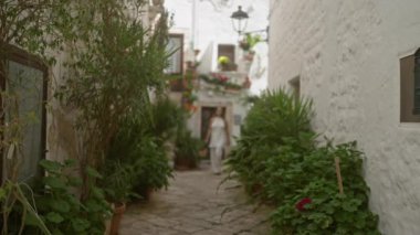 Genç, İspanyol bir kadın locorotondo, puglia, italya 'nın büyüleyici beyaz sokaklarını keşfediyor, güneşli bir günde yemyeşil bitkiler, çiçekler ve eski kasaba mimarisi ile çevrili..