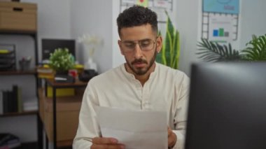 Arka planda çeşitli bitkiler ve organizasyonel malzemeler olan bir ofiste evrak okuyan genç bir İspanyol..