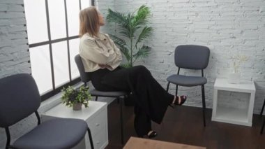 Genç, çekici, sarışın, kadın, modern, kapalı, beyaz tuğlalı duvarları, saksısı bitkileri ve minimalist mobilyaları olan bir bekleme odasında bekliyor..