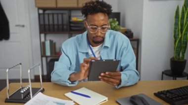 Ofiste çalışan Afro-Amerikalı genç bir adam çalışma masasında not alırken tablet kullanmaya odaklanmış..