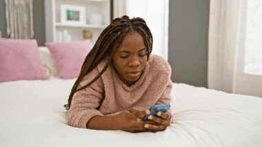 Evde saçı örgülü düşünceli bir Afrikalı kadın akıllı telefonunu sıradan bir yatak odasında karıştırıyor..