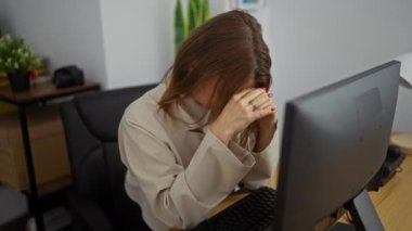 Ofis ortamında stresli bir kadın, sarı saçları ellerinde dinleniyor, ofis malzemeleriyle çevrili ve bilgisayar üzerinde çalışıyor..