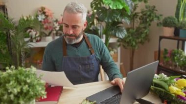 Laptop 'ta çiçek dükkanı işleten yakışıklı sakallı bir adam. Evrakları ve renkli çiçekleri olan bir ev..