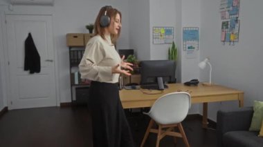 Kulaklık takan genç, çekici, sarışın bir kadın modern bir işyerinde el kol hareketleri yapıyor. İçeride, organize raflar ve masa ekipmanlarıyla..