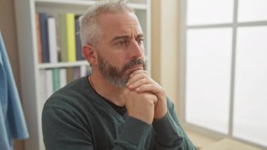 Sakallı, düşünceli, yetişkin, beyaz bir adam. Kapalı bir yerde oturuyor. İyi aydınlatılmış bir ev ortamında düşünceli görünüyor..