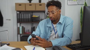 Sakallı, gözlüklü, masasında akıllı bir telefonla modern ofis ortamında çalışan Afrikalı genç bir Amerikalı..