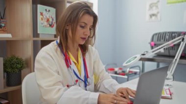 Veteriner kliniğinde dizüstü bilgisayarda çalışan genç sarışın bir veteriner steteskop takıyor ve arkasında tıbbi kitaplar ve ekipmanlar var..