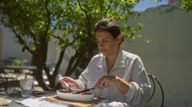 Genç bir İspanyol kadın, İtalya 'da bir açık hava restoranının terasında yemek yiyor. Gölge ağaçlarının altında oturuyor ve sakin bir yemek atmosferinde yemeğine odaklanıyor..
