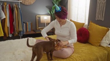 Kafkasyalı yetişkin bir kadın yatak odasında bir yatakta oturuyor, parti şapkası takıyor ve köpeğiyle iletişim kuruyor..