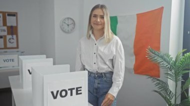 Sarışın, beyaz bir kadın, İrlanda 'daki kapalı bir üniversite odasında gülümsüyor ve arka planda İrlanda bayrağı olan oy verme kabinlerinin yanında başparmağını kaldırıyor..