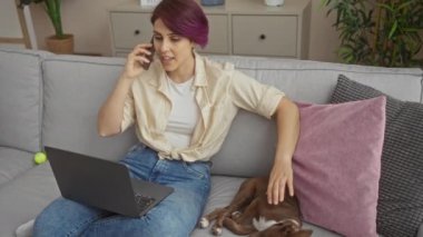 Evde dizüstü bilgisayarla çalışan, rahat bir oturma odasında köpeğini okşayan, renkli saçlı genç bir kadın..