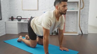 Genç adam spor salonunun içinde arka planda ağırlıkları olan mavi bir paspas üzerinde esneme egzersizleri yapıyor..