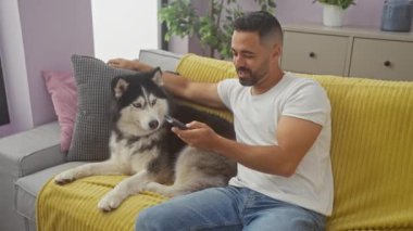 Mutlu İspanyol bir adam, günlük giysiler içinde evcil köpeğiyle sarı bir kanepede, uzaktan kumandalı, rahat bir odada..