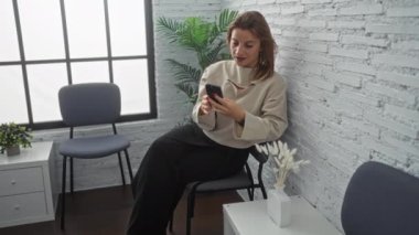 Beyaz bir kadın kapalı bir bekleme odasında oturmuş telefonuna gülümsüyor. Etrafı modern dekorlar, bitkiler ve minimalist mobilyalarla çevrili..