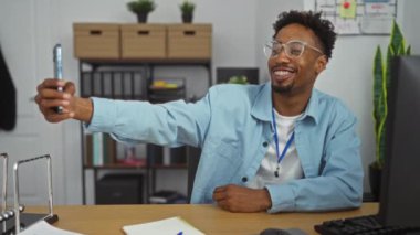Sakallı, mavi gömlekli, selfie çeken ve modern ofis ortamında gülümseyen Afrikalı genç bir Amerikalı..