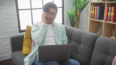 Genç Asyalı bir adam birden fazla iş yapıyor, telefonla konuşuyor ve gri kanepesi ve beyaz tuğlalı duvarı olan modern bir oturma odasında dizüstü bilgisayar kullanıyor..