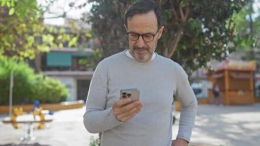 Günlük süveter giyen olgun bir adam, güneşli bir parkta, kentsel geçmişe sahip bir akıllı telefon kullanır..