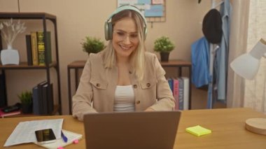 Evdeki ofisinde, kulaklık takan ve video görüşmesi sırasında gülümseyen genç sarışın bir kadın bilgisayarında bitkiler, kitaplar ve akıllı telefonuyla bir masada oturuyor..
