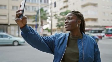 Saçları örgülü gülümseyen Afrikalı bir kadın şehir sokaklarında selfie çekiyor, kentsel yaşam tarzı ve teknoloji kullanımı sergiliyor