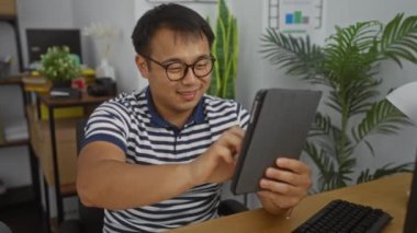 Ofisindeki yakışıklı Çinli adam bir video konferansı düzenliyor. Etrafı bitkiler ve ofis dekorlarıyla çevrili modern bir iş ortamında dijital bir toplantıya katılıyor..