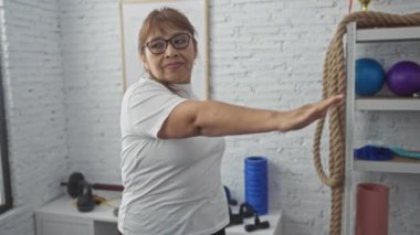 Orta yaşlı bir kadın, spor salonunda egzersiz yapıyor, beyaz bir arka planı var, kollarını egzersiz ekipmanları ve iplerinin yanında beyaz bir tişörtle açıyor..