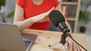 Kırmızı bluzlu beyaz bir kadın modern bir stüdyo ortamında mikrofon ve dizüstü bilgisayarlı bir podcast kaydediyor..