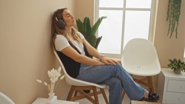 Genç ve çekici sarışın bir kadın kulaklık takıyor, kapalı bir bekleme odasında bitkilerle dinleniyor, arka planda penceresi olan bir sandalyeye oturuyor ve kapalı alanda bitkiler yetiştiriyor..