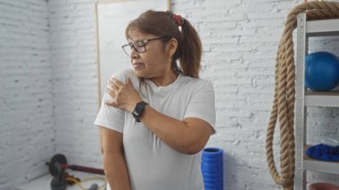 Spor salonundaki orta yaşlı İspanyol bir kadın omzuna dokunuyor. Etrafında egzersiz ekipmanları var. İp ve ilaç topu da var..