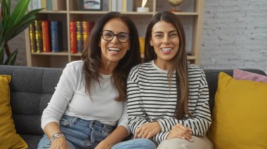 Gülümseyen bir anne ve kız, sıcak bir oturma odasındaki kanepeye yakın oturarak aile sevgisini ve arkadaşlığını sembolize eder..