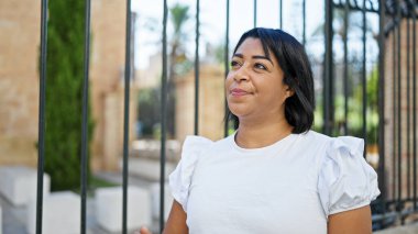 Beyaz tişörtlü orta yaşlı İspanyol kadın yeşil şehir parkında gülümsüyor.