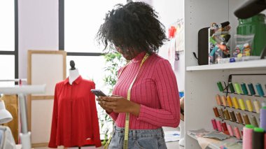 Kumaş ve mankenli renkli bir terzi dükkanında akıllı telefon kullanan Afrikalı Amerikalı bir kadın..