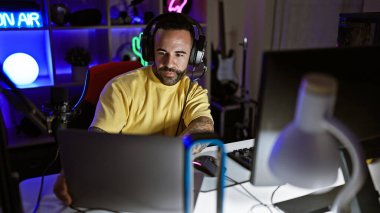 Karanlık bir oyun odasında sakallı genç bir İspanyol adam. Kulaklıkları, mikrofonu ve renkli neon ışıkları var..