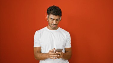 Kırmızı arka planda akıllı telefon ve kulaklık kullanan sakallı yakışıklı bir İspanyol.