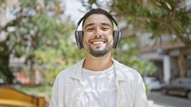Gülümseyen sakallı, kulaklık takan ve beyaz gömlekli genç bir adam açık havada, yemyeşil bir şehir parkında müzik dinlemekten hoşlanıyor..