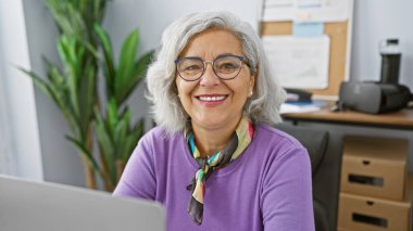 Gri saçlı, gözlüklü ve ofiste eşarp takan gülümseyen olgun bir kadın.