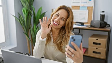 Ofiste akıllı telefonuyla video görüşmesinde gülümseyen genç İspanyol kadın el sallıyor.