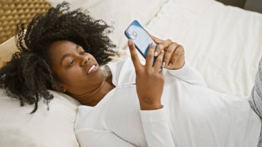 Afro-Amerikalı bir kadın, sıcak, iyi ışıklandırılmış bir yatak odasında elinde akıllı bir telefonla, kapalı bir yatakta dinleniyor..