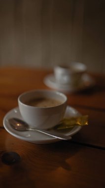 Beyaz bir fincanda kapuçinosu olan bir kafenin samimi bir ambiyansı, yanında da bir çay kaşığı ve ahşap bir masada bir şeker paketi..