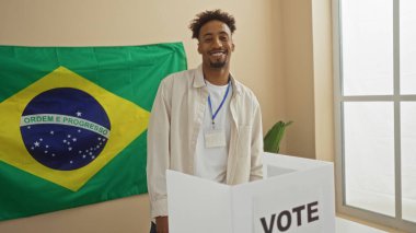 Sakallı Afrikalı genç bir Amerikalı oy odasında arka planda Brezilya bayrağı olan bir oy sandığının yanında duruyor..