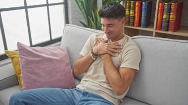 Yakışıklı İspanyol adam, elleri göğsünde gülümsüyor, rahat bir oturma odasında gri bir koltukta oturuyor..