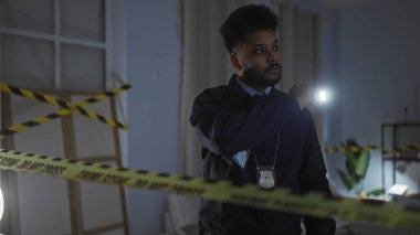 Afro-Amerikalı bir adam ön planda dikkat bandıyla feneriyle aydınlatılmış bir suç mahallini evin içinde inceliyor..