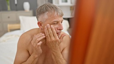Düşünceli orta yaşlı bir adam yatakta otururken yüzüne dokunuyor. Sabah rutinini ya da yatak odasında cilt bakımını tasvir ediyor..